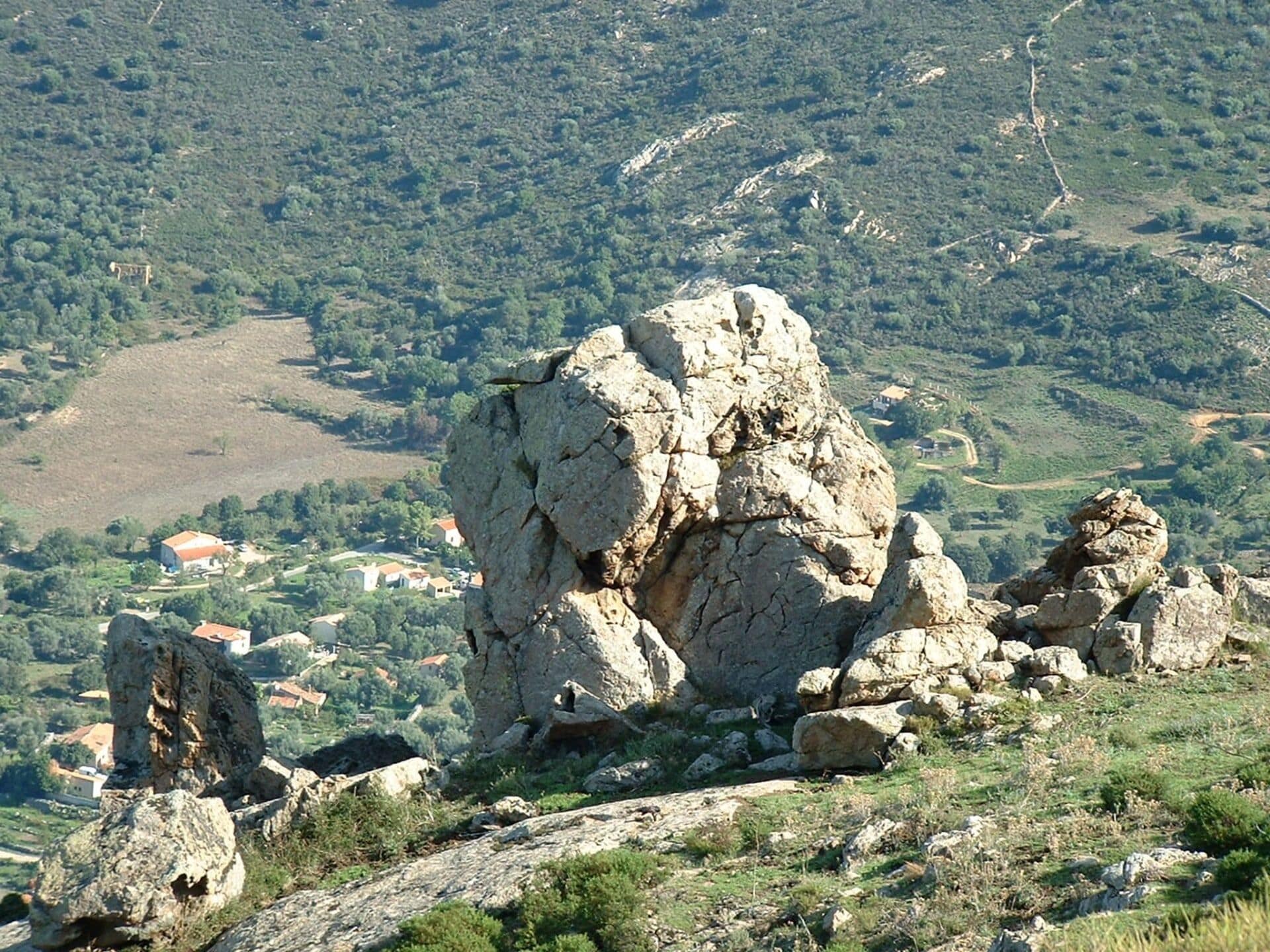 Eine zerklüftete Landschaft mit einer markanten Felsformation im Vordergrund und einem Tal mit verstreuten Gebäuden im Hintergrund.