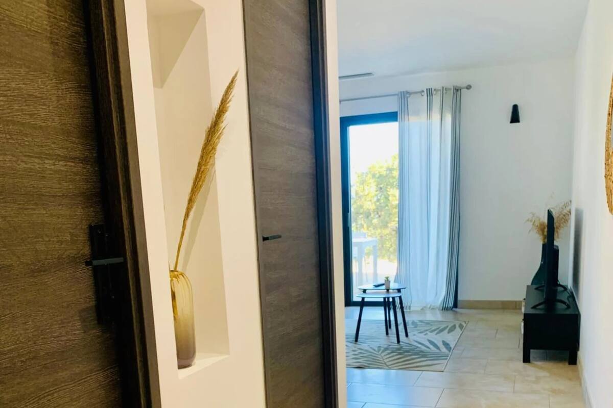 A hallway with a dark wooden door, a white wall with a decorative element, and a tiled floor leading to a room with a window and curtains.