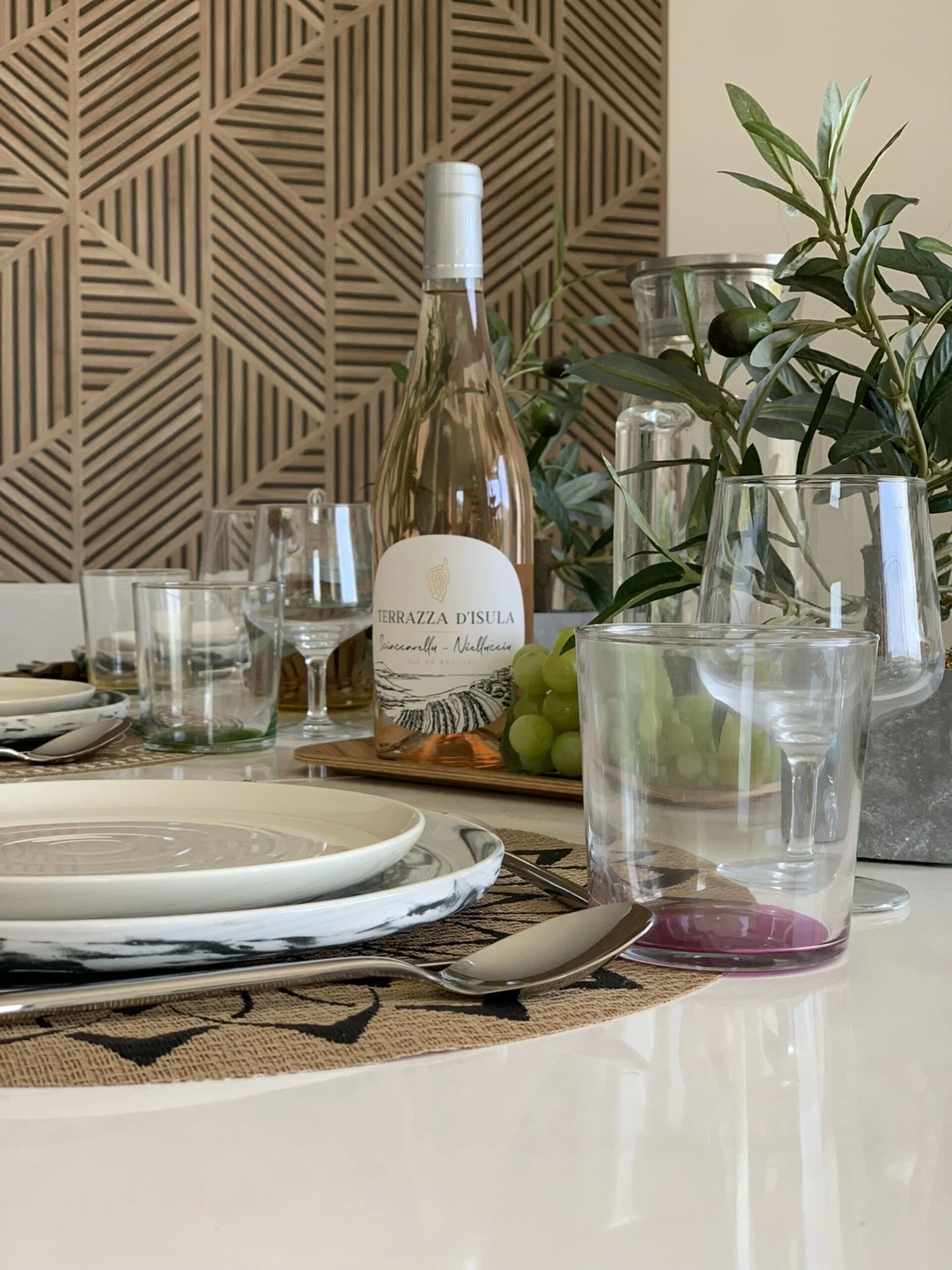 Una mesa dispuesta con platos, cubiertos, una botella de vino, copas y una planta.