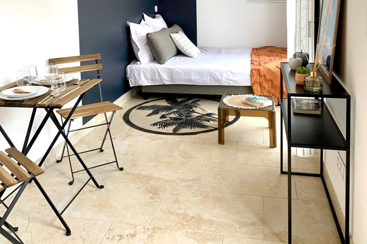 A compact room with a bed, a dining table with chairs, a small side table, and a wall-mounted air conditioner.
