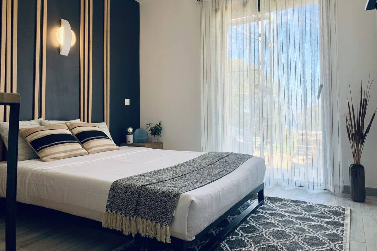 A modern bedroom with a bed, a bedside table, a lamp, and a window with sheer curtains.
