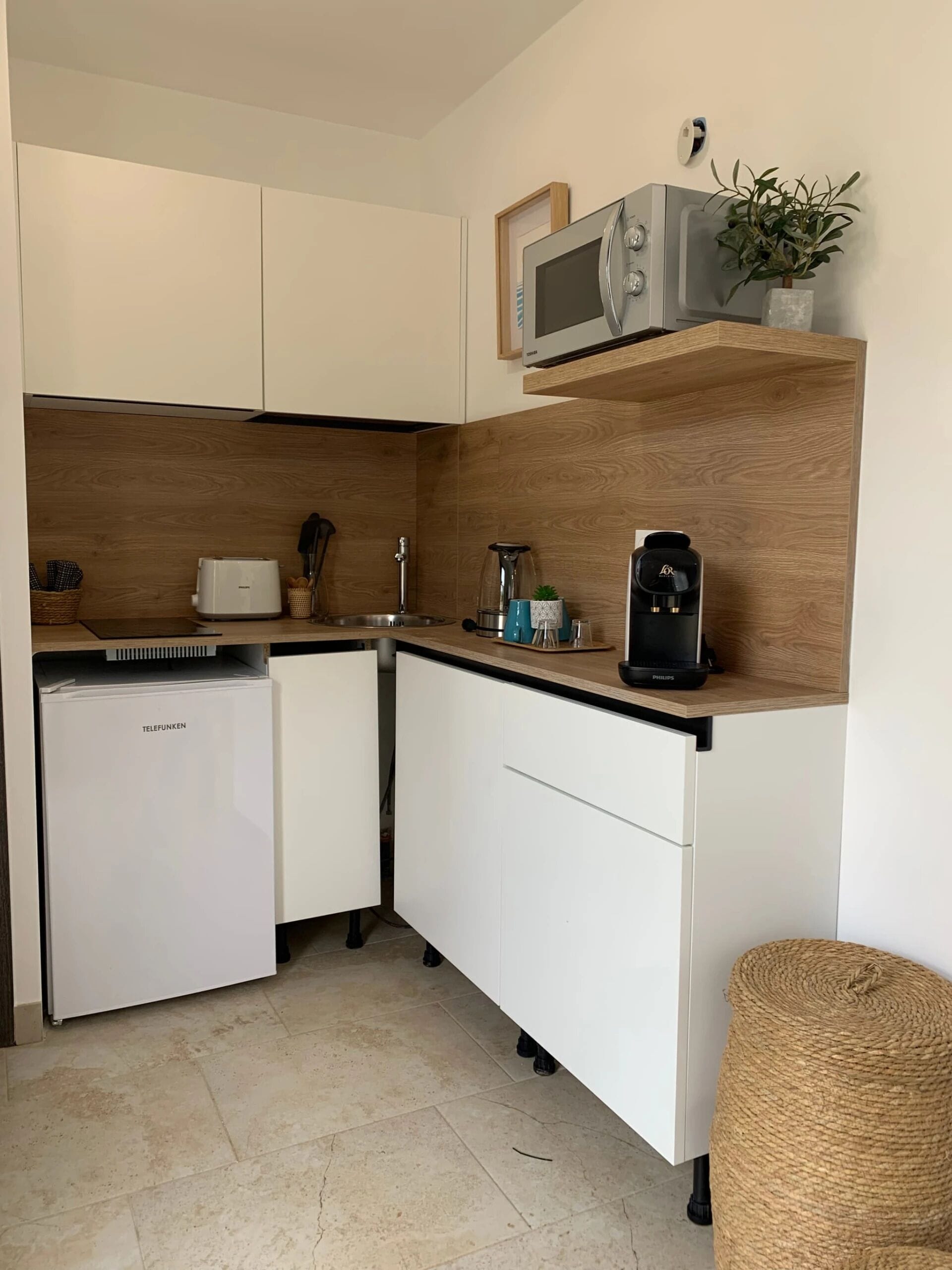 A compact kitchen with white cabinets, a built-in microwave, a small refrigerator, and a coffee machine.