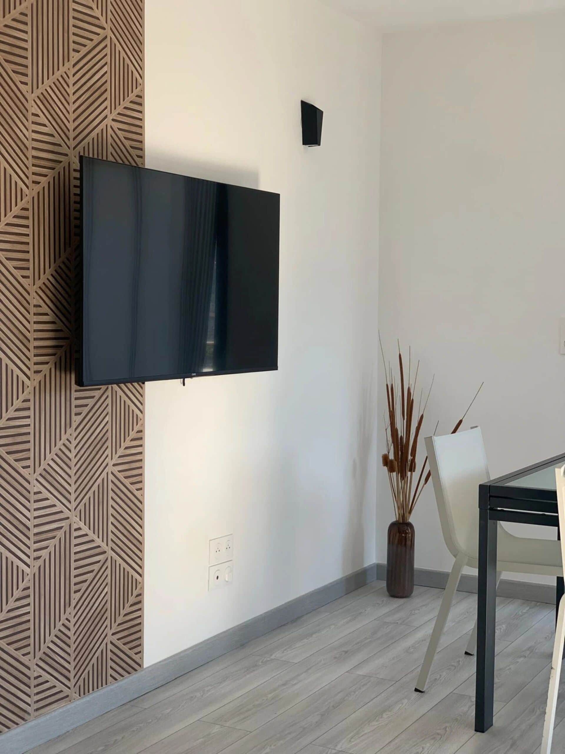 A modern living room with a flat-screen TV mounted on the wall, a patterned wall panel, a white chair, and a small table.