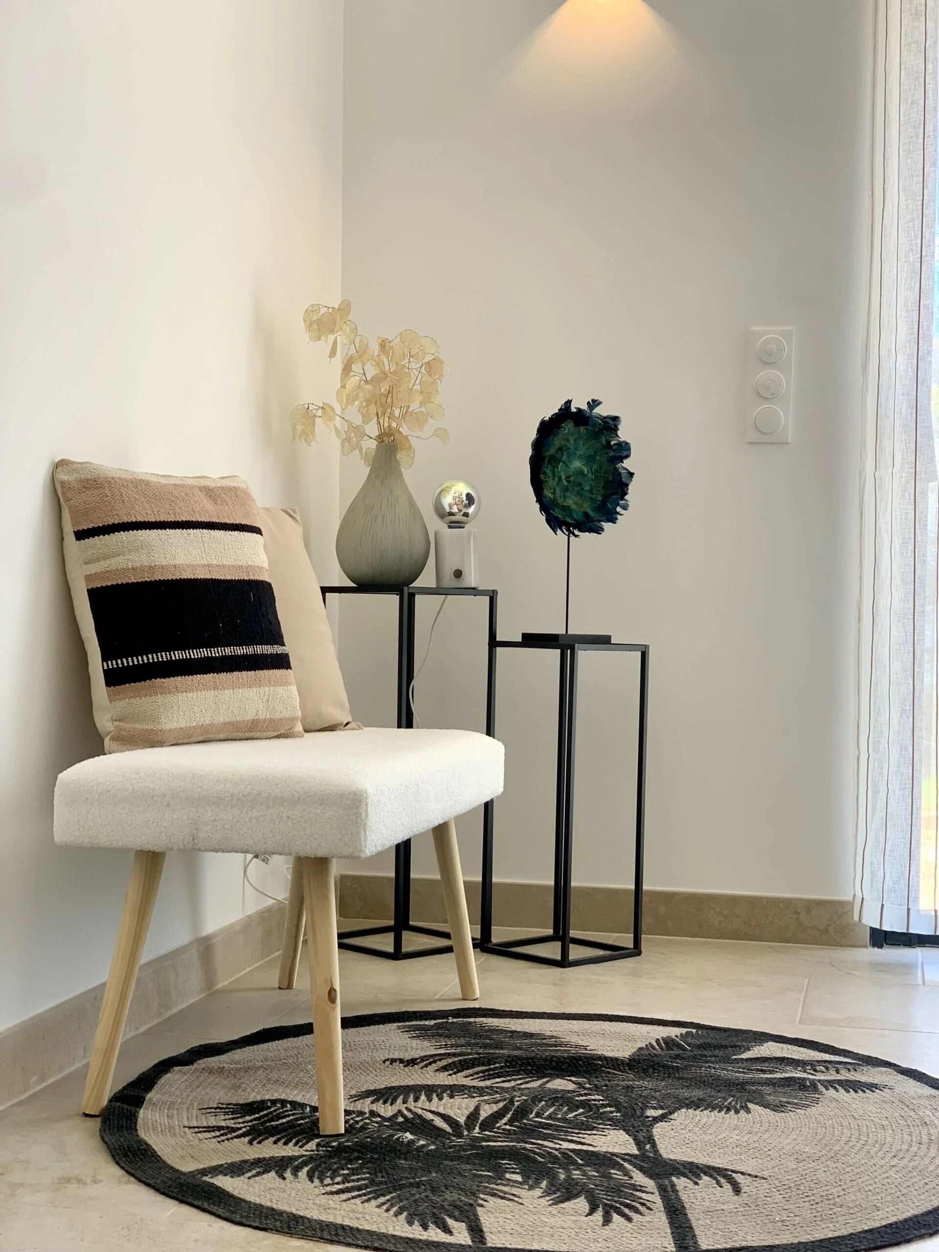 Una silla con un cojín a rayas, un florero con flores secas, una esfera decorativa y una mesita con un tapete redondo con motivos de palmeras.