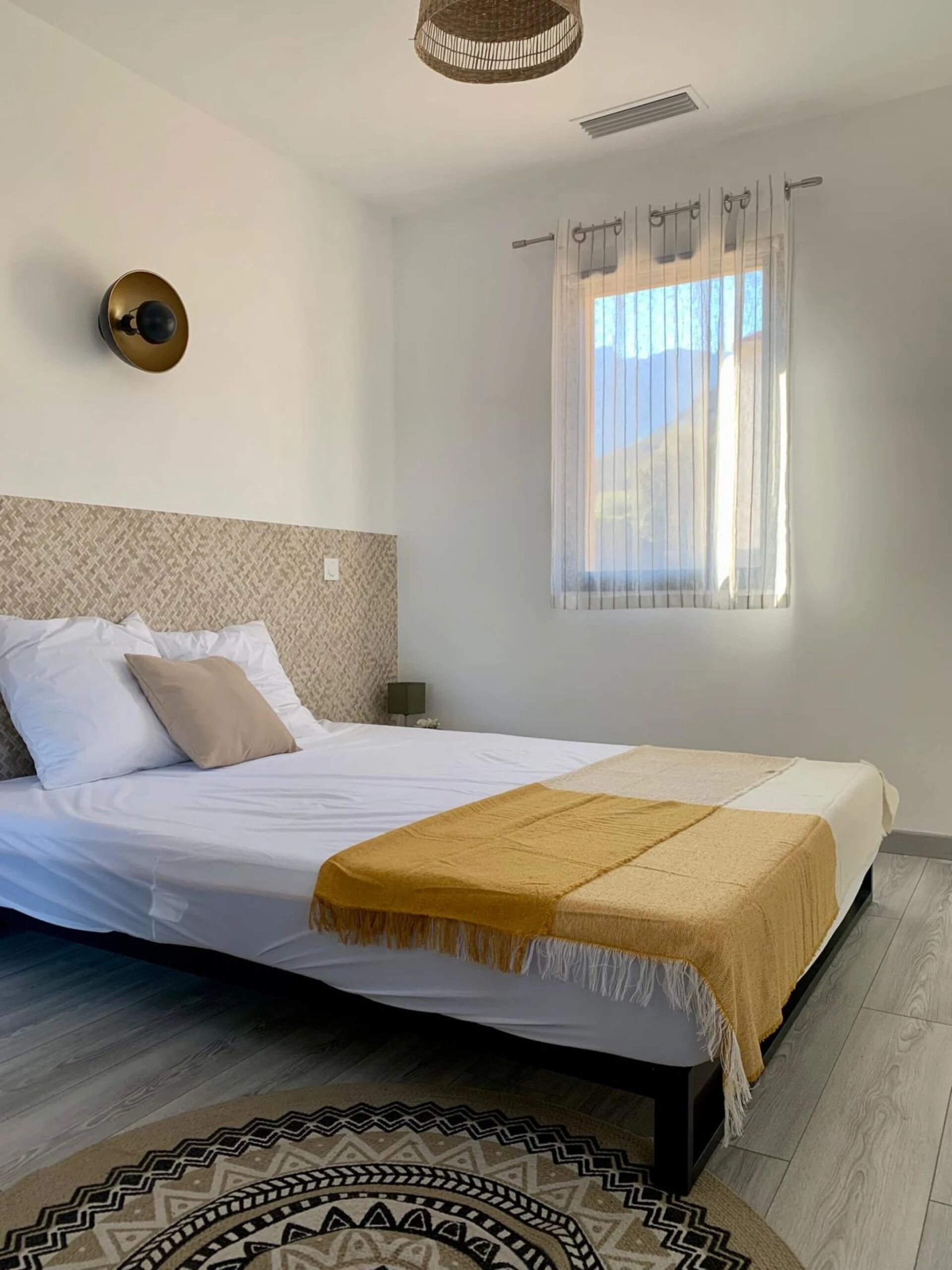 Een netjes ingerichte slaapkamer met een bed, een bedlampje, een raam met gordijnen en een kamerkleed met dessin.