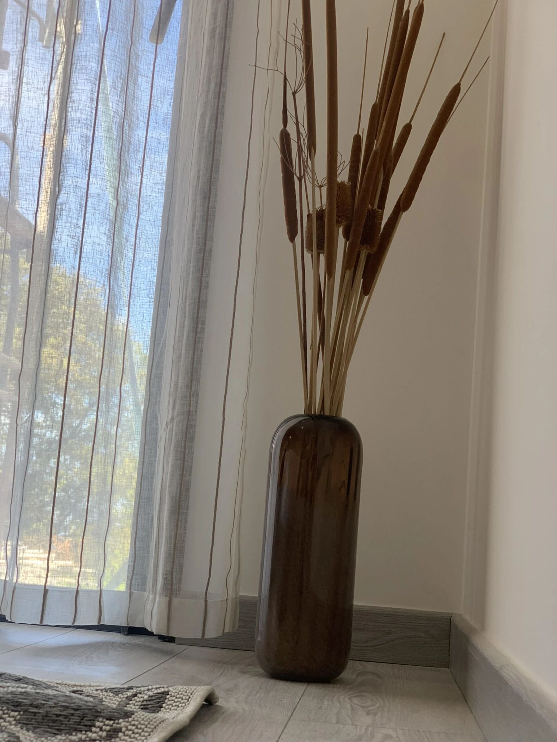 A room with a large window, a wooden floor, and a vase with dried plants.
