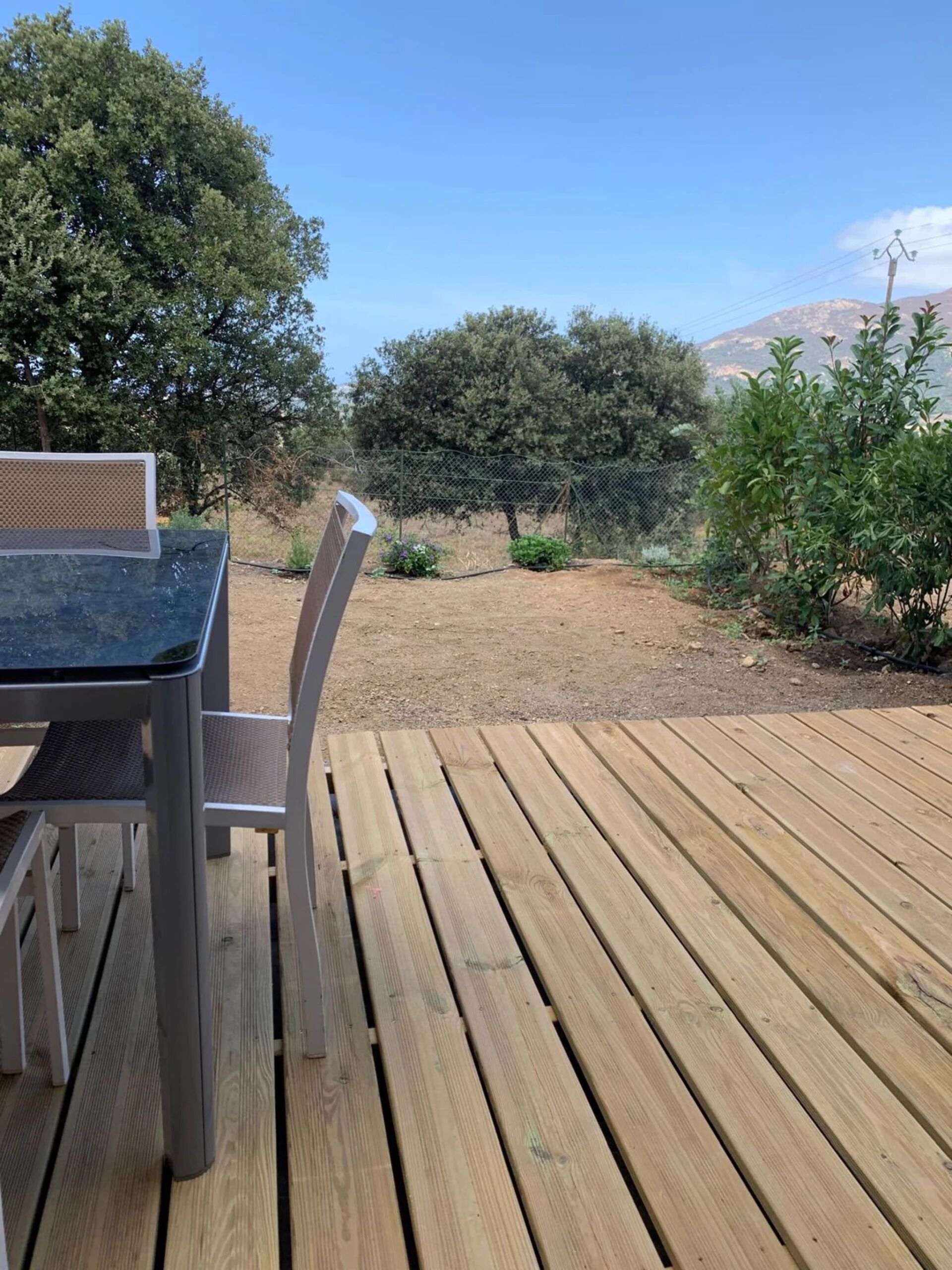 A wooden deck with a table and chairs, surrounded by greenery and a fence.