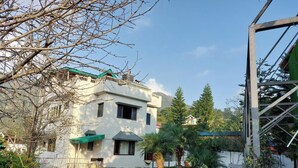 A two-story building with a green roof, surrounded by a well-maintained garden with hedges and palm trees. - Alinda House Naukuchiatal by THE GHAUR (Uttarakhand)