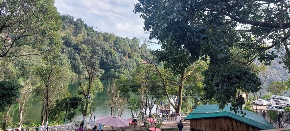 Restaurante à beira do lago com mesas ao ar livre, cercado por vegetação exuberante e colinas.