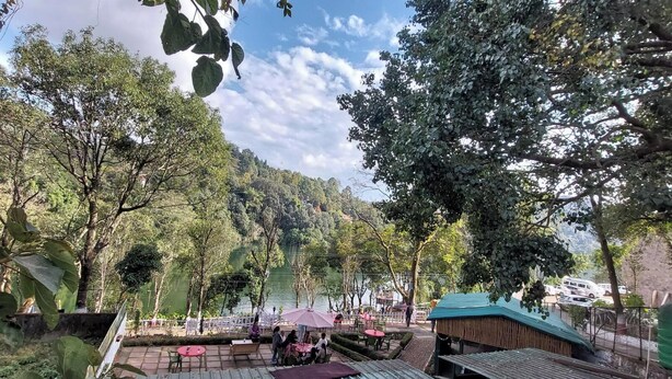 A lakeside restaurant with outdoor seating, surrounded by trees and a view of the lake. - Alinda House Naukuchiatal by THE GHAUR (Uttarakhand)