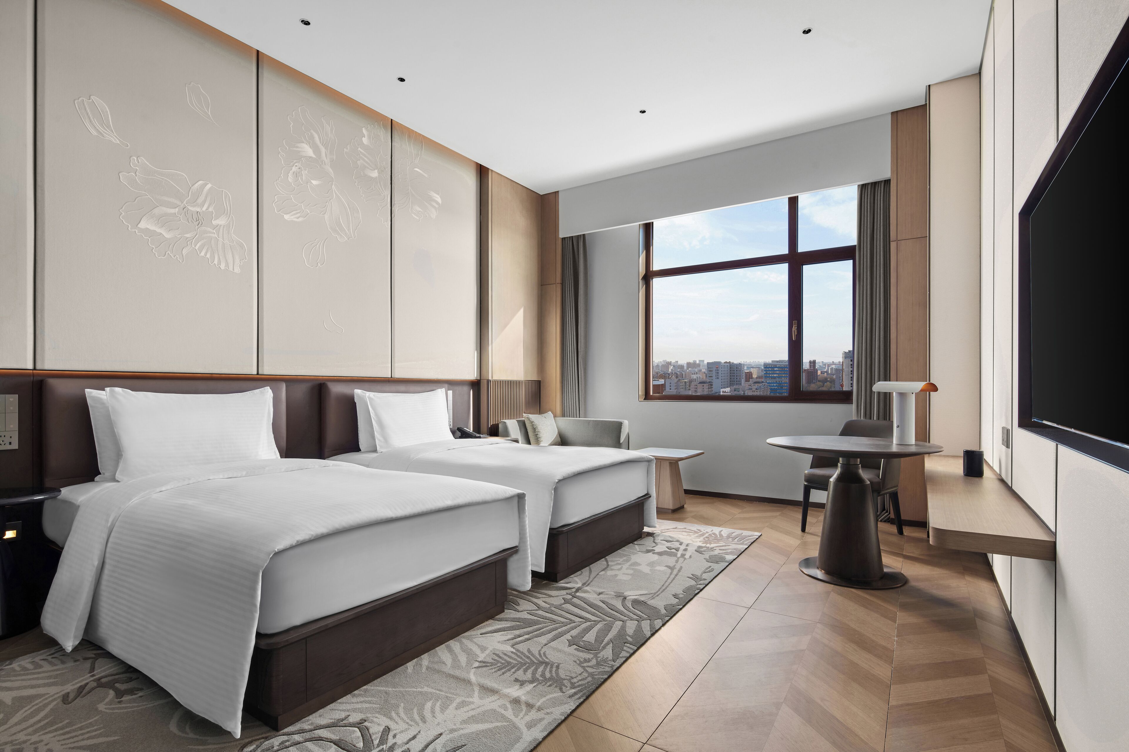 Habitación de hotel con dos camas, un escritorio, una silla, un televisor y un ventanal con vistas a la ciudad.