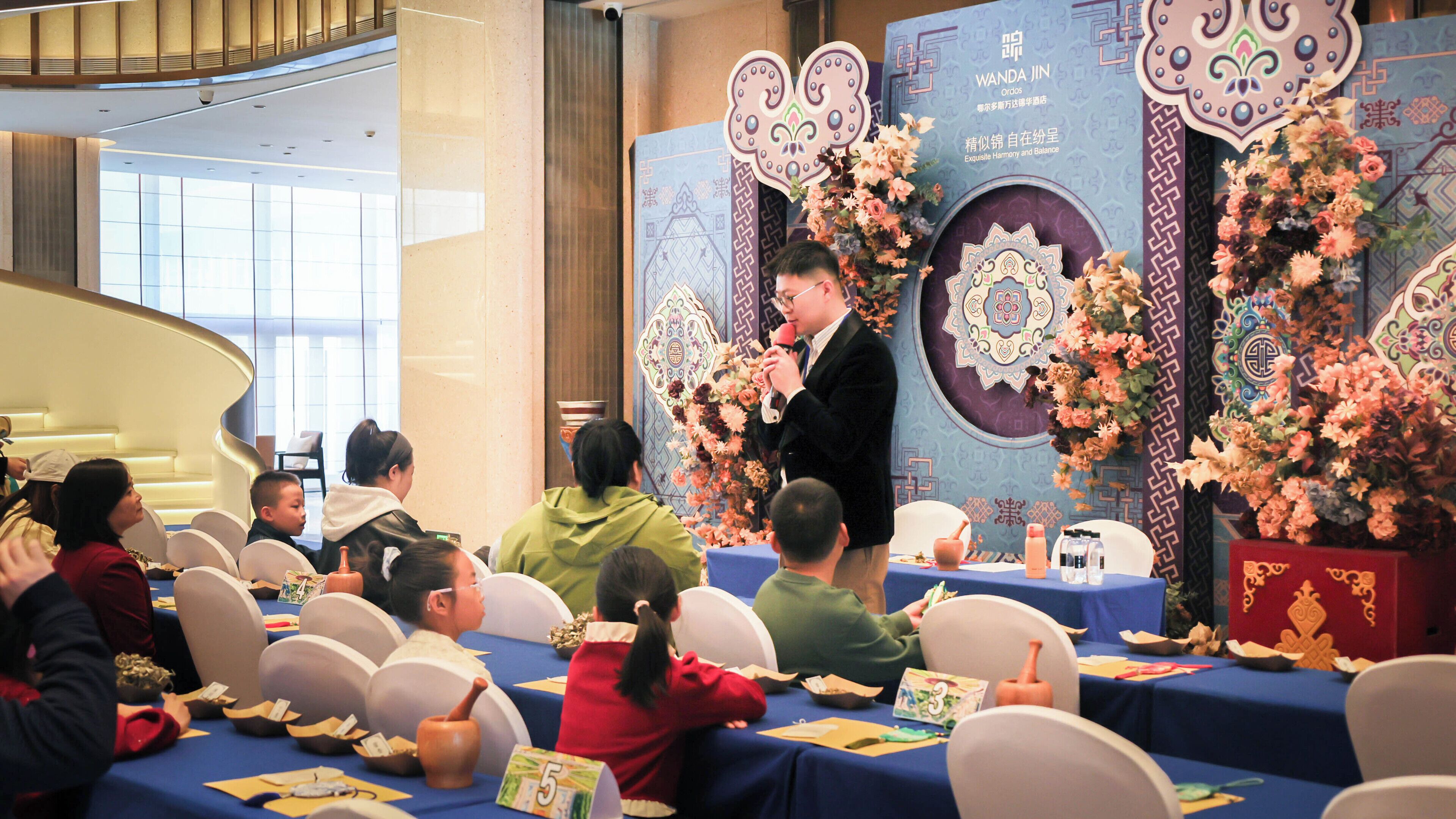 A conference room with a speaker at a podium, audience seated at tables, and decorative floral arrangements.