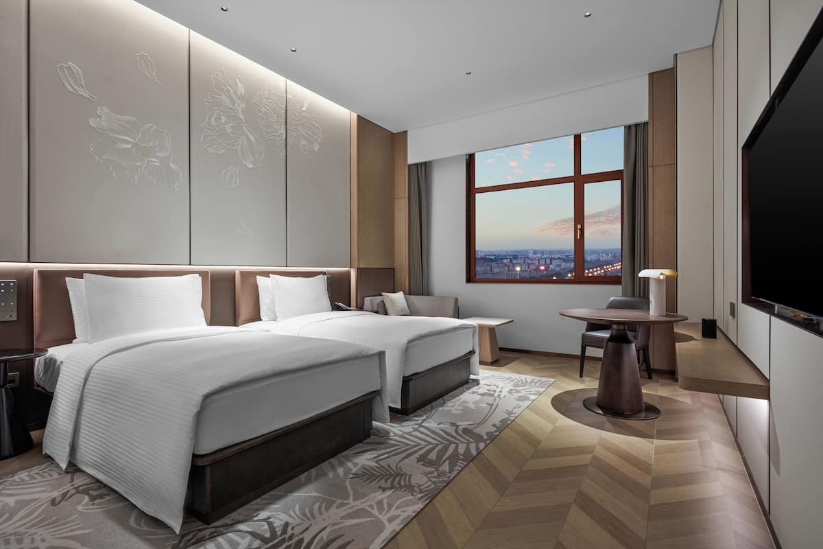 A modern hotel room with two beds, a large window with a city view, a small table, and a TV.
