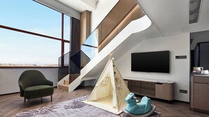 Un salon moderne avec une grande fenêtre, un fauteuil vert, un espace de jeu aménagé comme une tente, un cheval à bascule bleu et un escalier.
