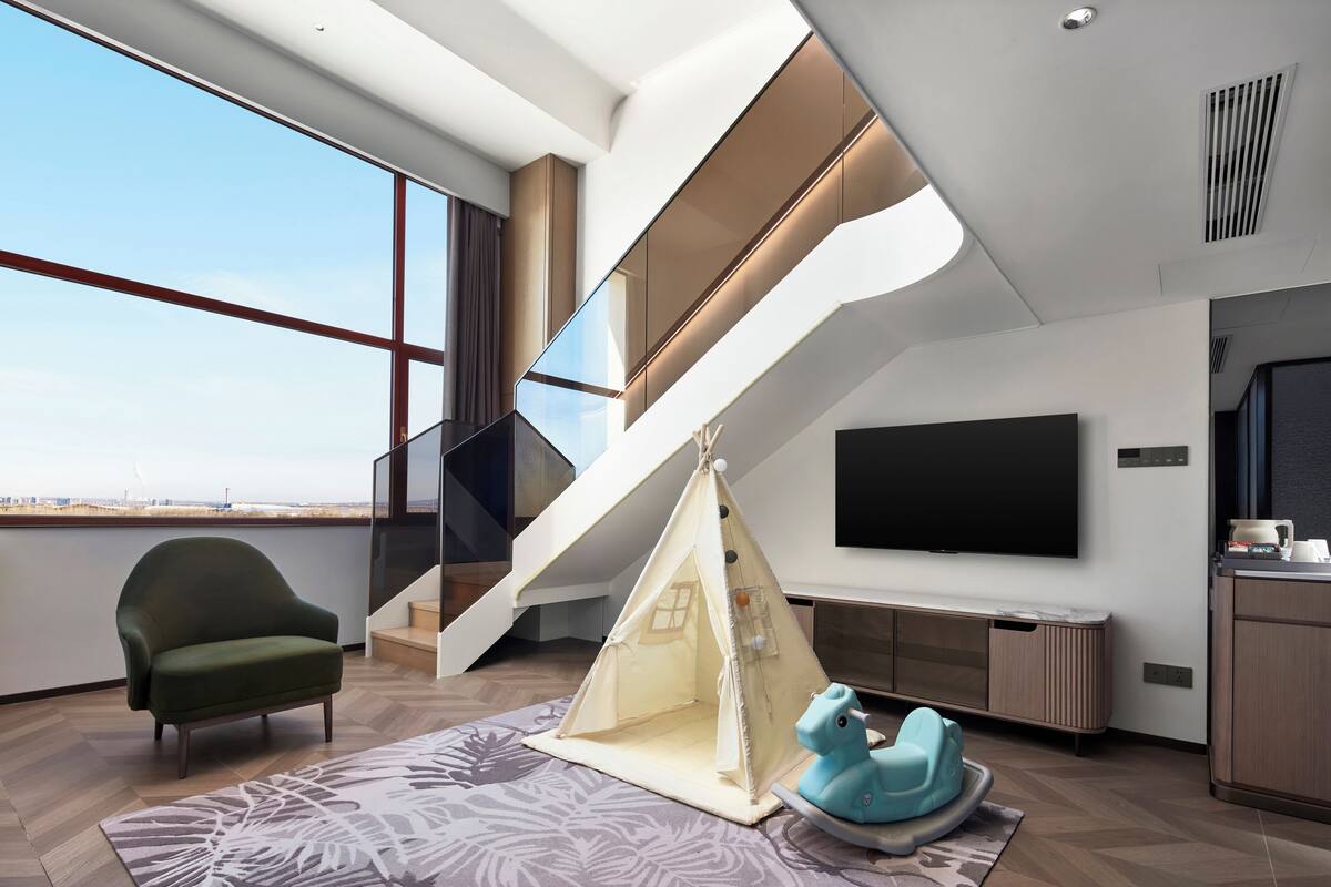 A modern living room with a large window, a green armchair, a tent-like play area, a blue rocking horse, and a staircase.