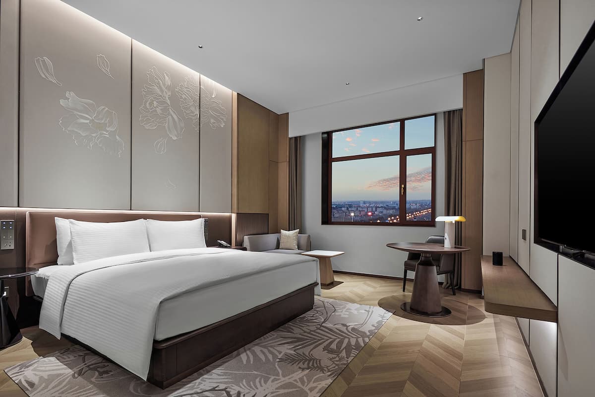 A modern hotel bedroom with a large bed, a desk, a chair, and a TV.