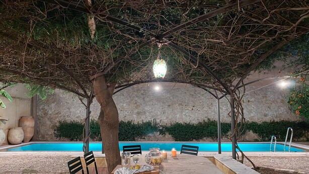 Un espace extérieur aménagé pour les repas, avec une table dressée pour deux personnes, entourée de chaises, sous un arbre agrémenté d’un luminaire suspendu. On aperçoit une piscine à l’arrière-plan.