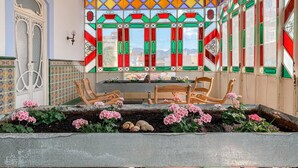 Un balcon offrant une vue sur la ville, agrémenté de vitraux colorés, d’un coin salon avec des chaises et d’un parterre fleuri où poussent des fleurs roses.