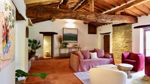 Ein rustikales Interieur mit freiliegenden Holzbalken, einer Steinwand, einem Sofa, einem Sessel und einem Landschaftsbild.