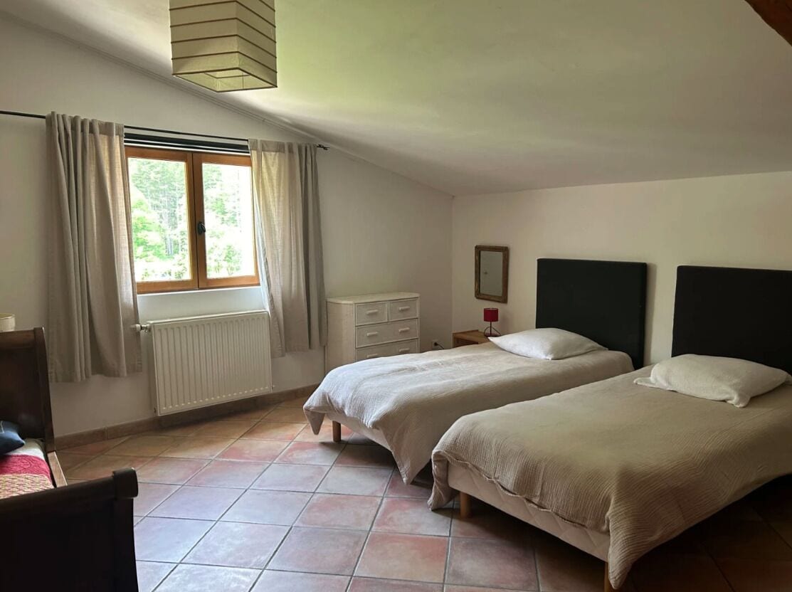 A bedroom with two beds, a window with curtains, a radiator, a nightstand, and a chair.