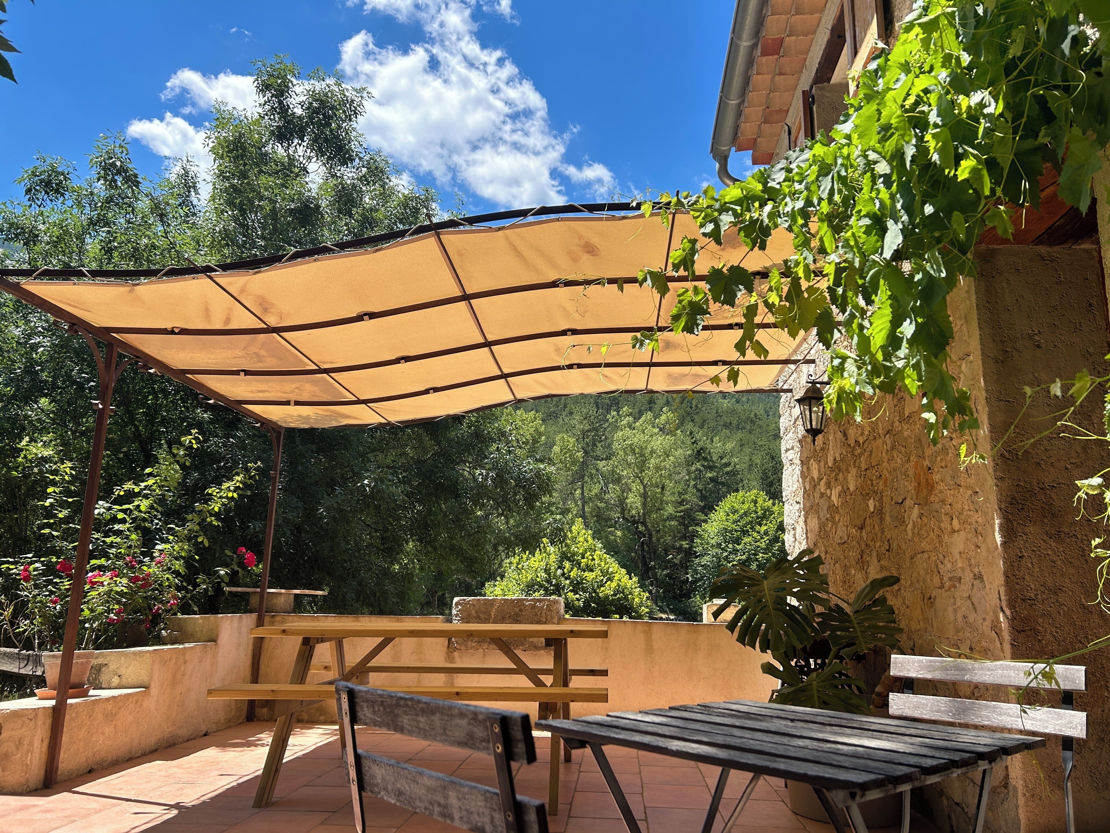 An outdoor patio with a shaded table and benches, surrounded by greenery and a stone wall.