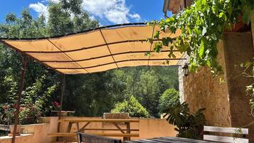 An outdoor patio with a shaded table and benches, surrounded by greenery and a stone wall.