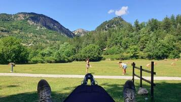 A person lying on the grass with a mountain view, a wooden fence, and hay bales in the background.