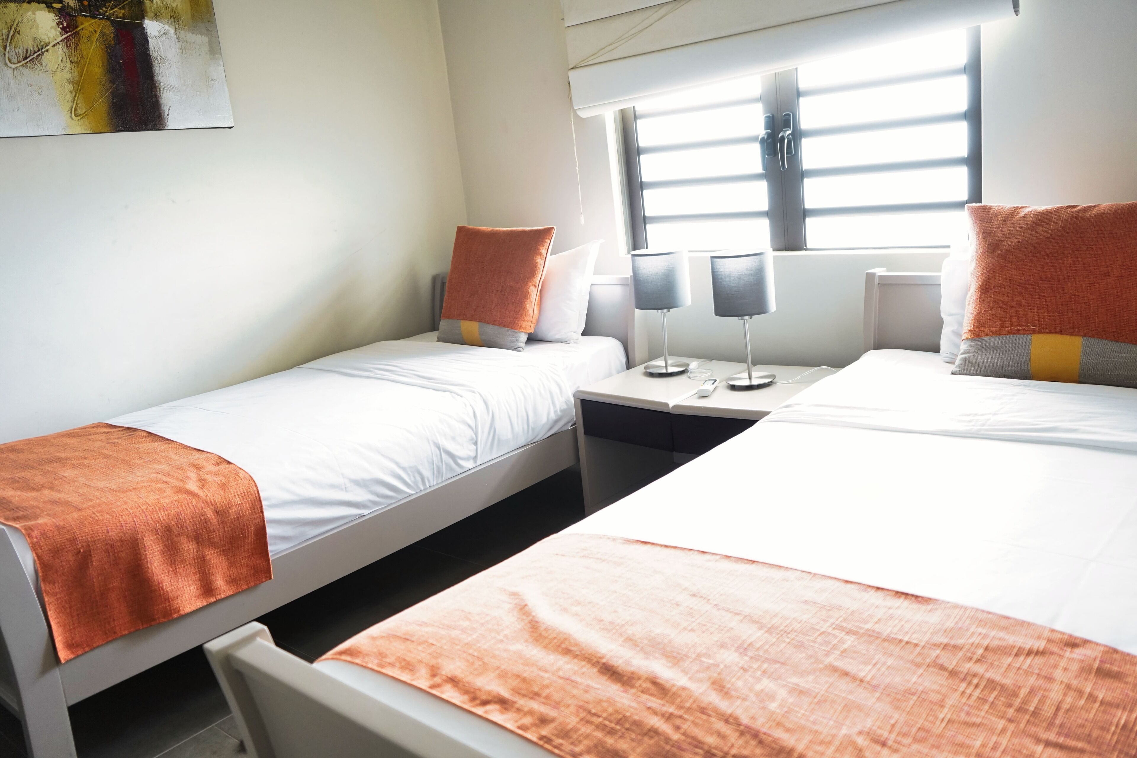 A hotel room with two single beds, each with an orange blanket, white bedding, and matching pillows. There are bedside tables with lamps and a window with blinds.