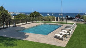 Una piscina con sillones de descanso, rodeada de vegetación y vista al mar con barcos.