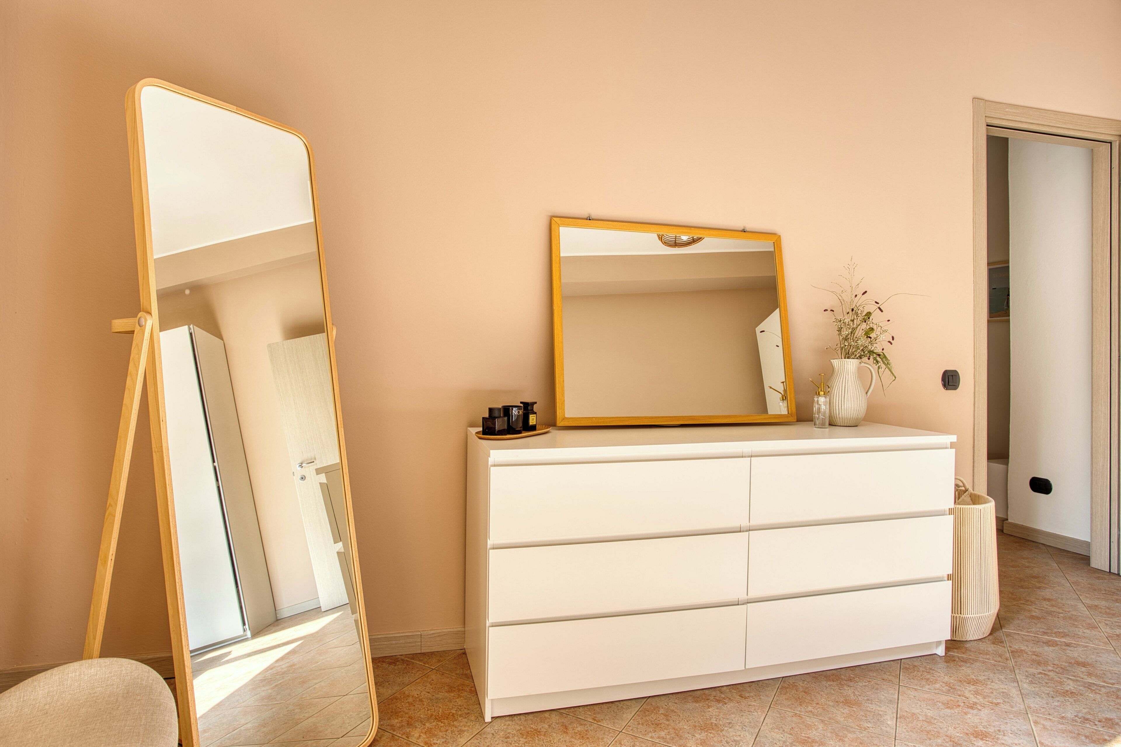 Une pièce avec une commode blanche, un miroir avec un cadre doré et une porte donnant sur une autre pièce.