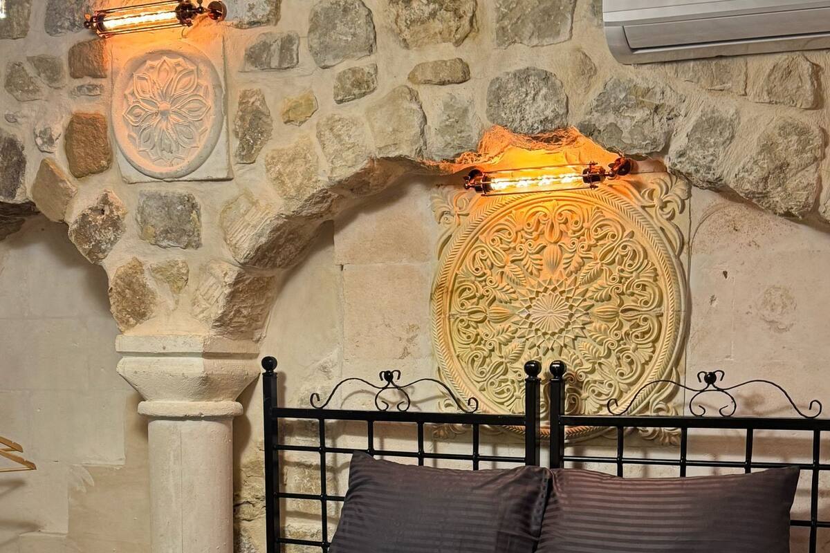 Ein Bett mit dunkler Bettwäsche, eine Steinwand mit geschnitzten Verzierungen und eine dekorative Leuchte darüber.