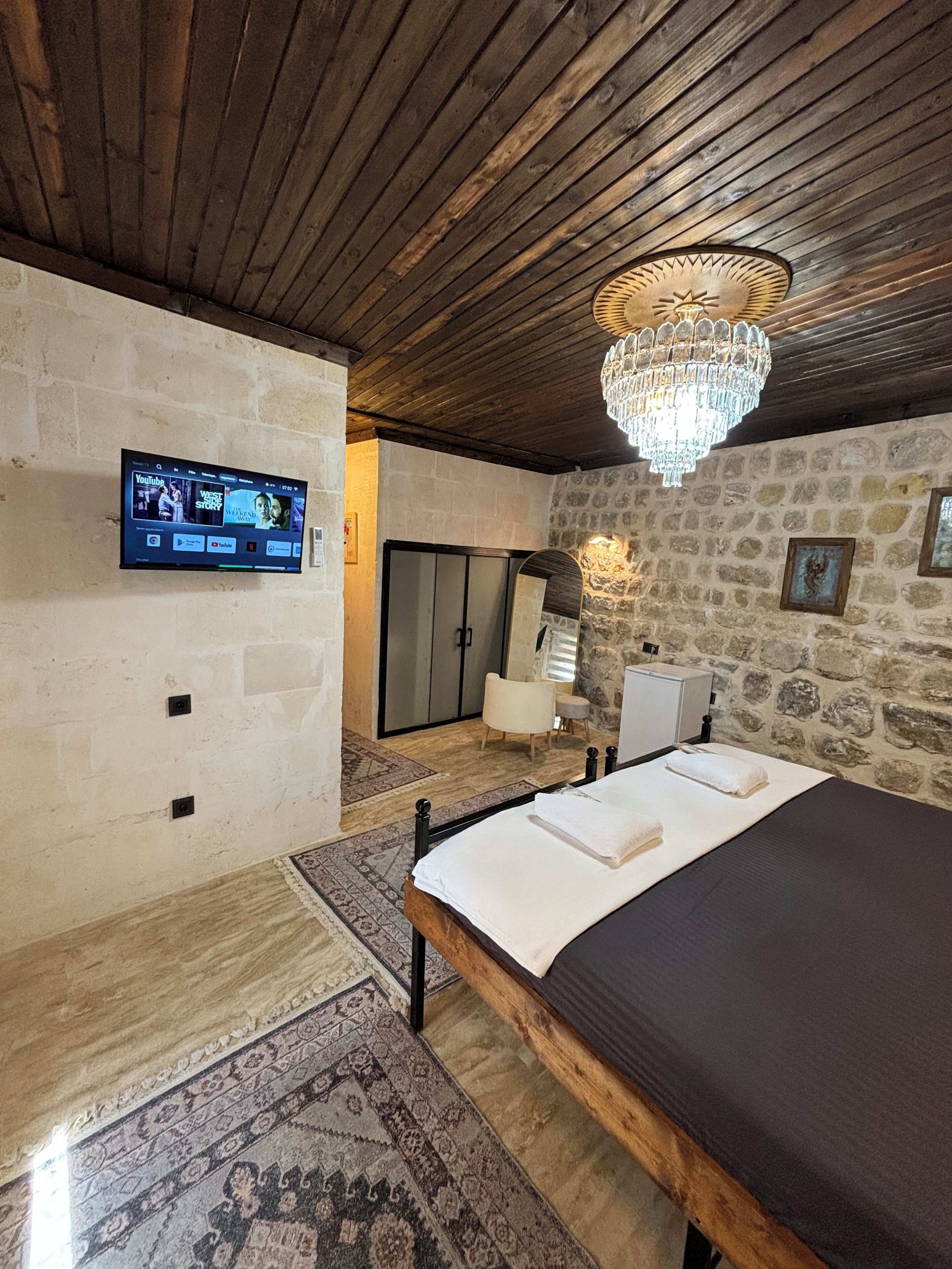 A bedroom with a bed, a television mounted on the wall, a chandelier, and stone walls.