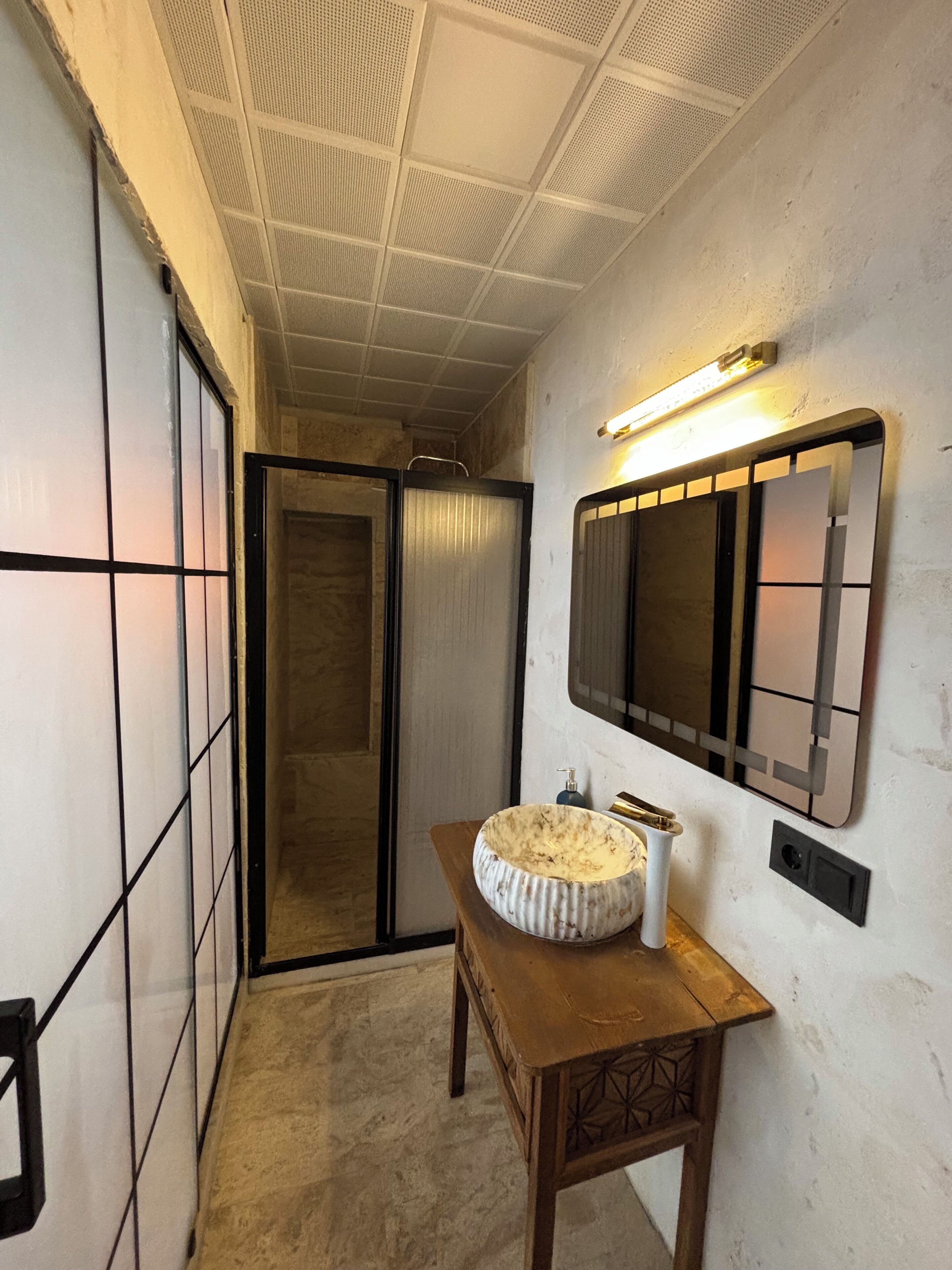 Une salle de bain avec un meuble-lavabo en bois, un lavabo rond et un miroir avec une lampe au-dessus.