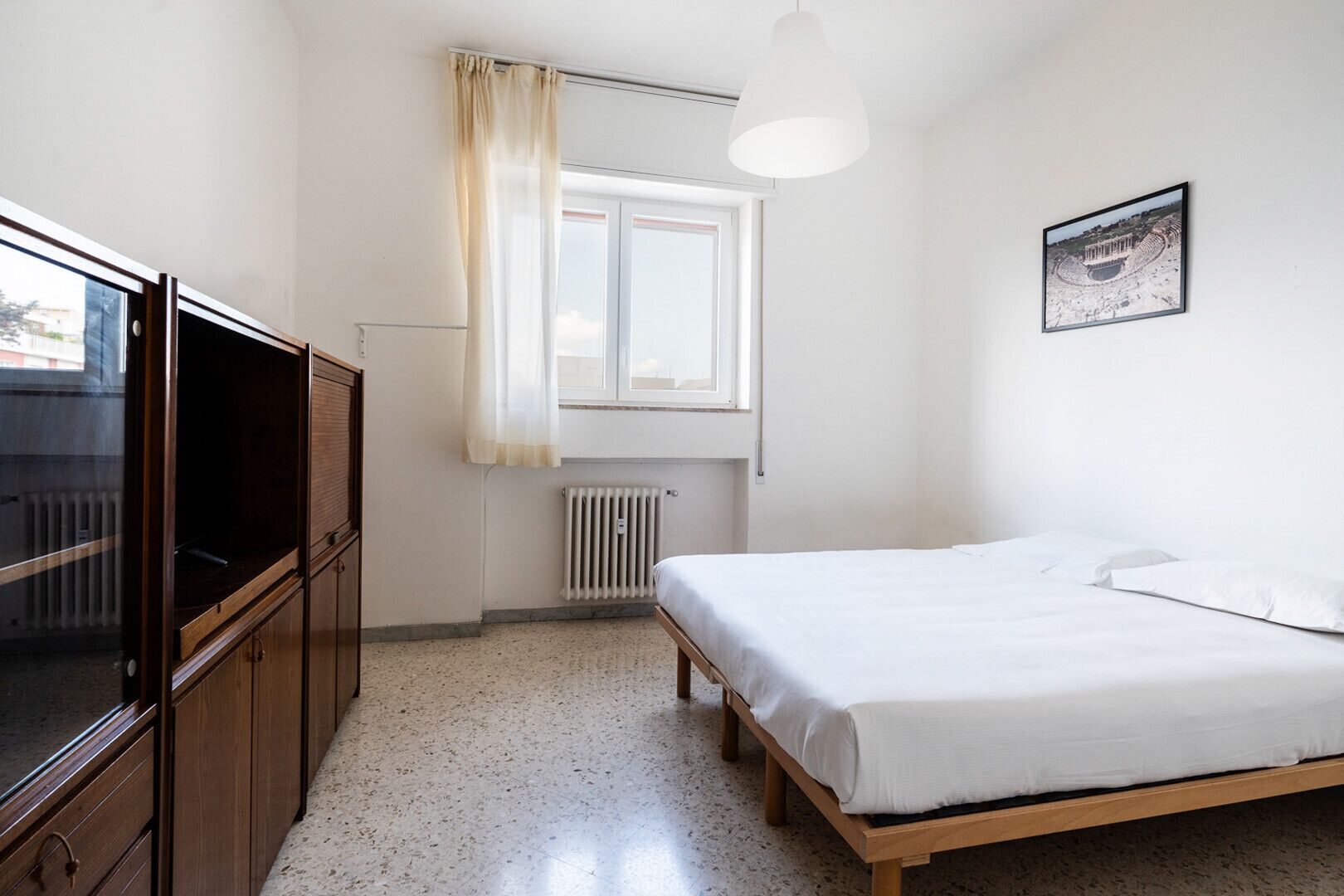 A bedroom with a bed, a window with curtains, a radiator, a wooden wardrobe, and a framed picture on the wall.