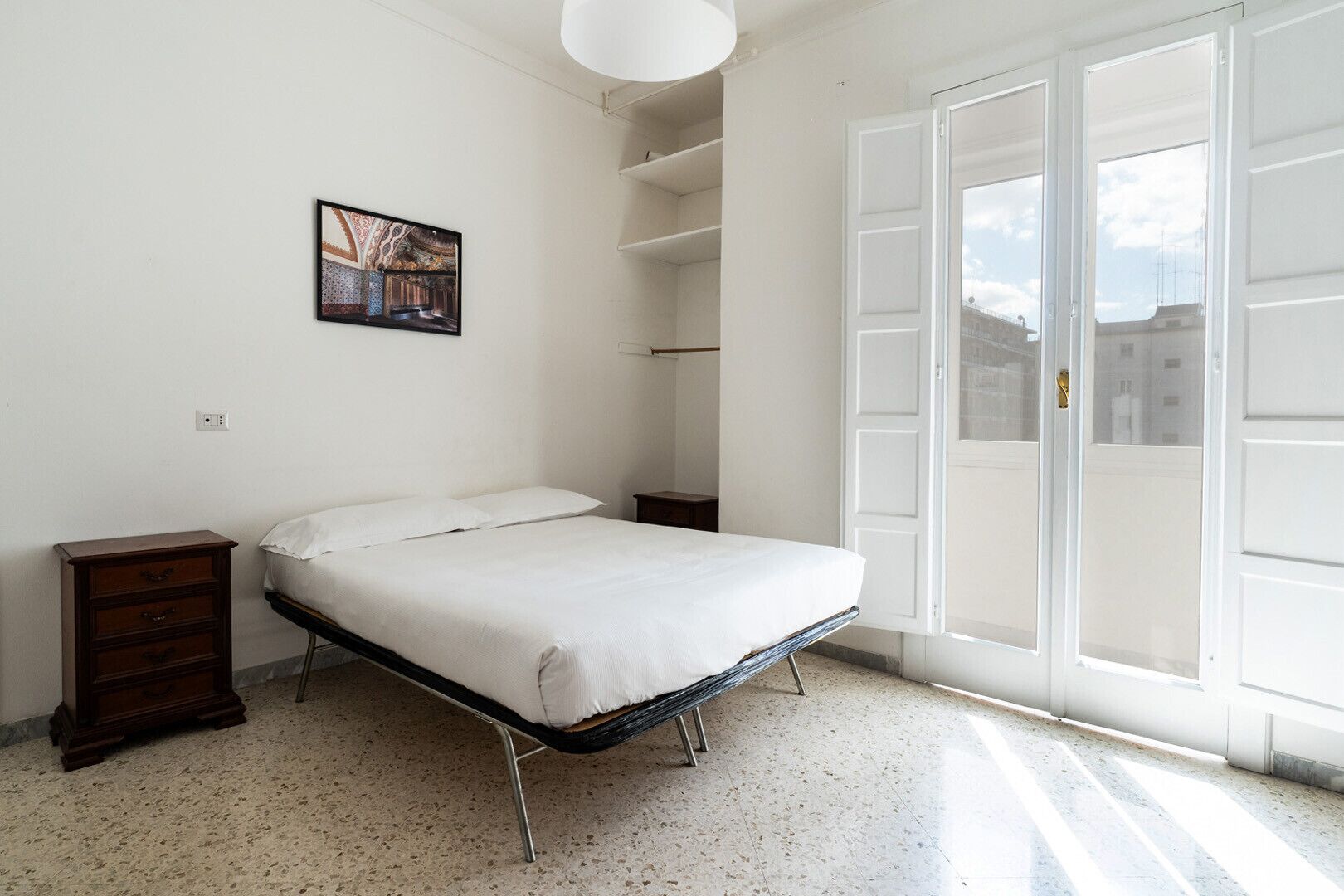 Un dormitorio con cama, cómoda, un cuadro y un amplio ventanal con vistas a edificios.