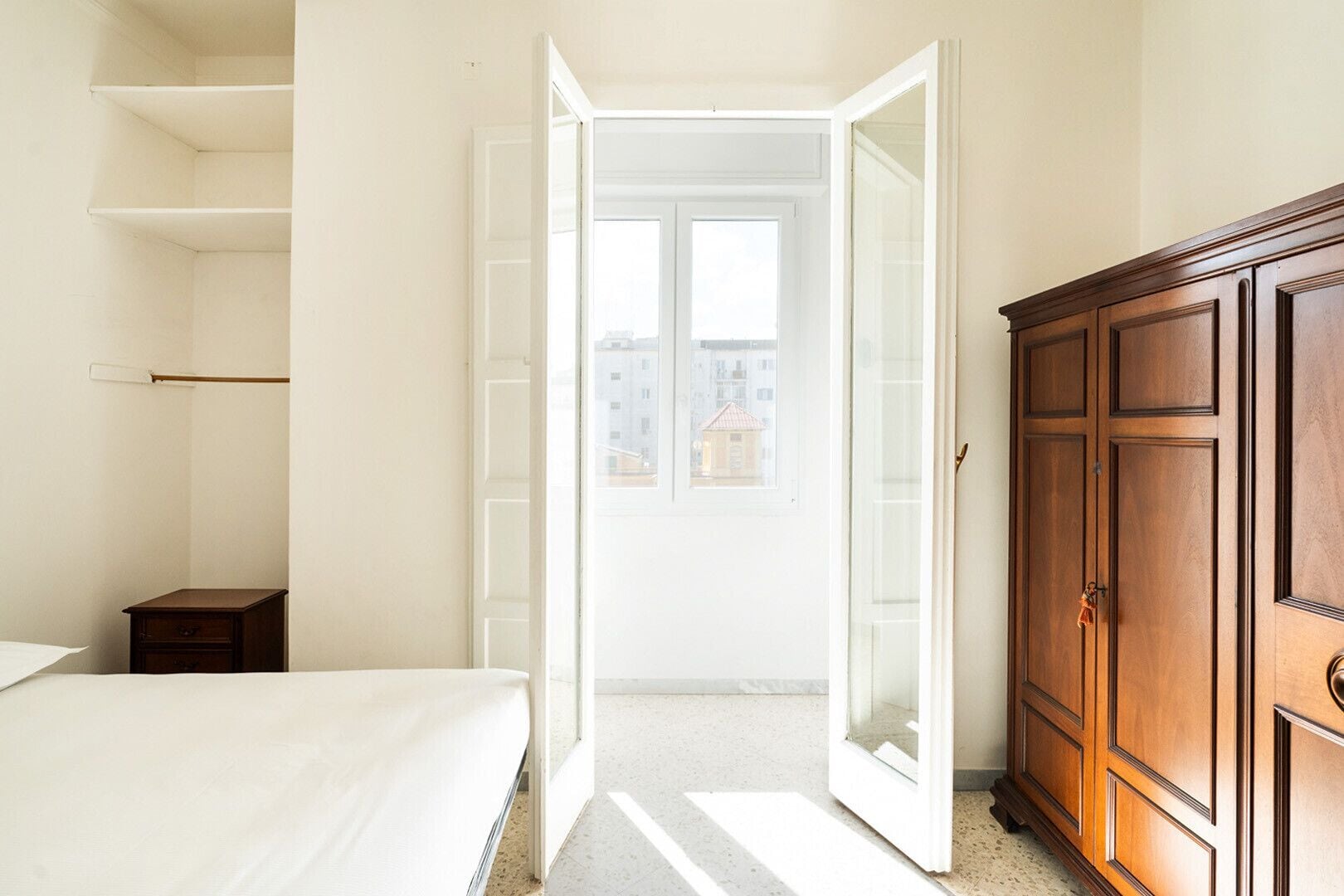 Quarto com cama, guarda-roupa de madeira e porta que dá para uma varanda com vista para edifícios.