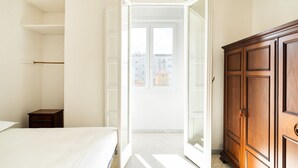 A bedroom with a bed, a wooden wardrobe, and a door leading to a balcony with a view of buildings.
