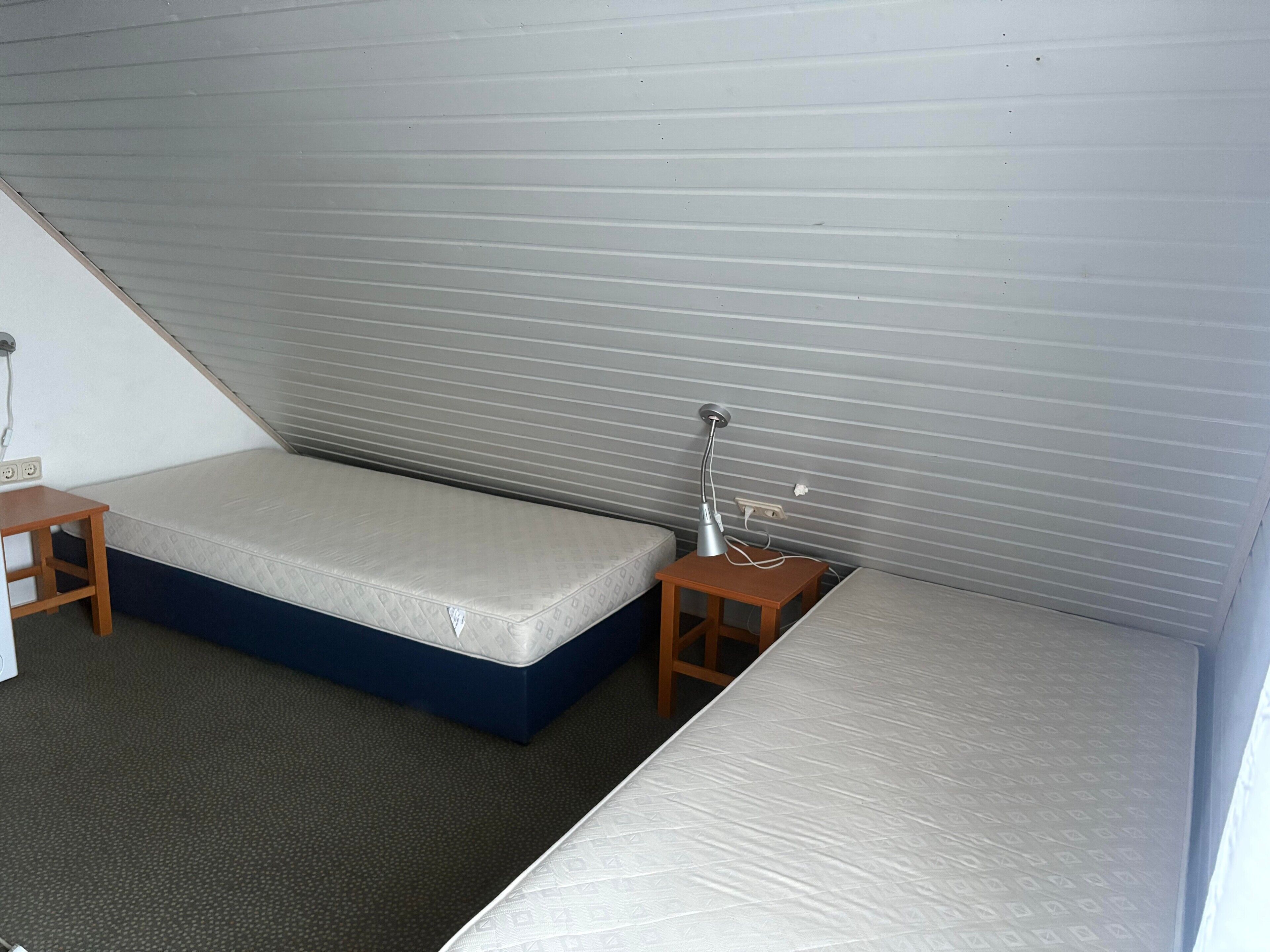 Een kleine kamer met enkel een bed, een nachtkastje en een lamp, met een schuine wand.