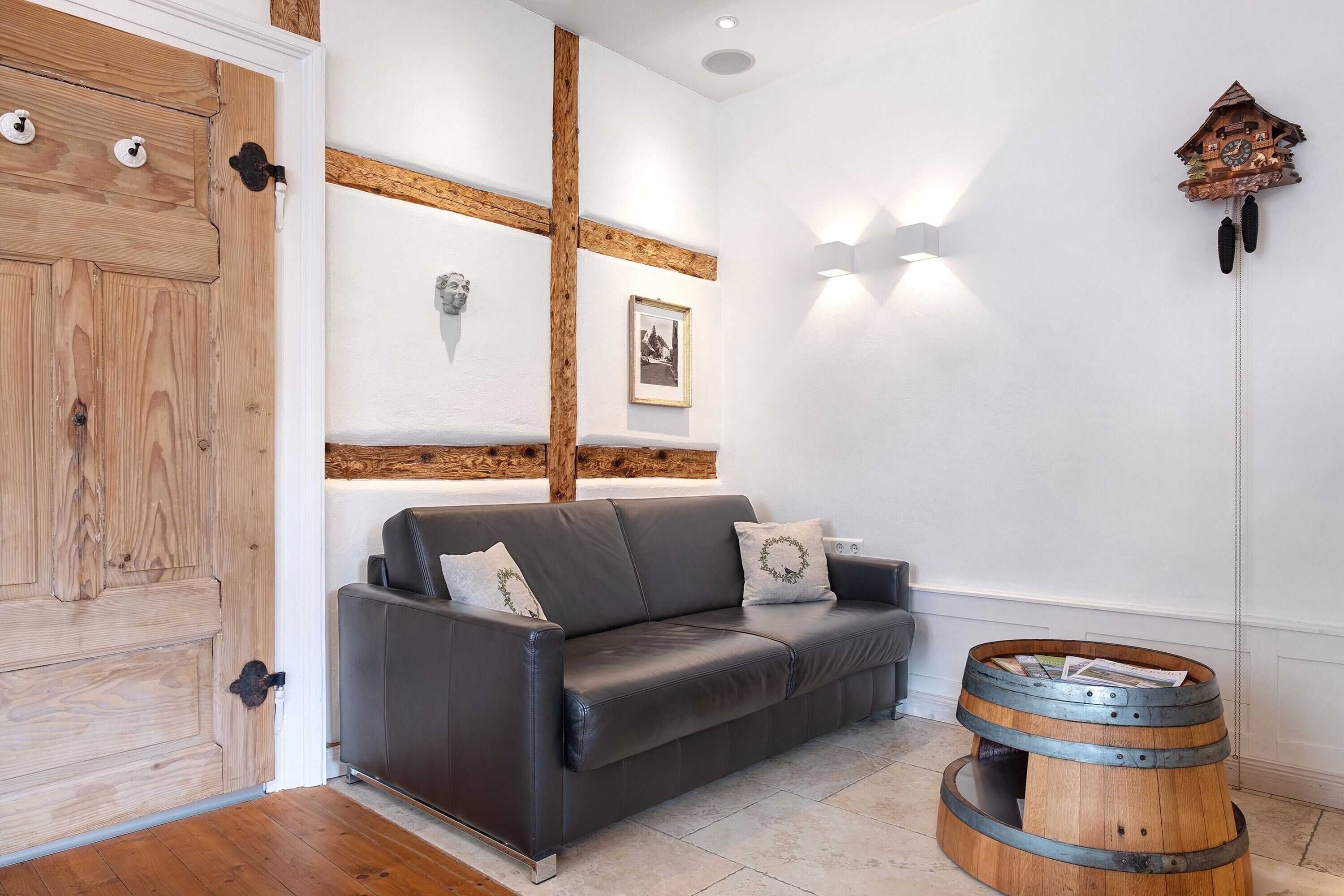 A room with a wooden door, a leather sofa, a wall-mounted light fixture, and a wooden barrel table.