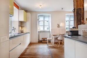 Une cuisine moderne avec des meubles blancs, un plancher en bois, un coin repas avec une table et des chaises blanches, et une grande fenêtre avec des stores.