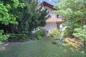 Una casa con un balcone, circondata da vegetazione rigogliosa e un piccolo prato con una sedia.