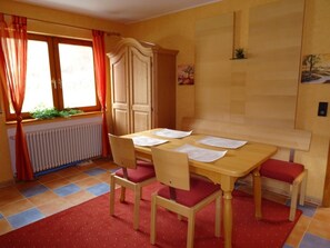 A dining room with a wooden table, four chairs, a radiator, a window with curtains, and a painting on the wall. - Apartment 'Mühlengrün' with Mountain View, Balcony and Wi-Fi (Oberwolfach)