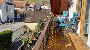 A balcony with a table and chairs, a flower box with plants, and a view of houses in the background. - Apartment 'Mühlengrün' with Mountain View, Balcony and Wi-Fi (Oberwolfach)