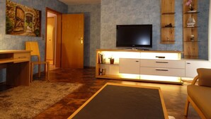 A living room with a flat-screen TV, wooden shelves, a desk, and a chair. - Apartment 'Mühlengrün' with Mountain View, Balcony and Wi-Fi (Oberwolfach)