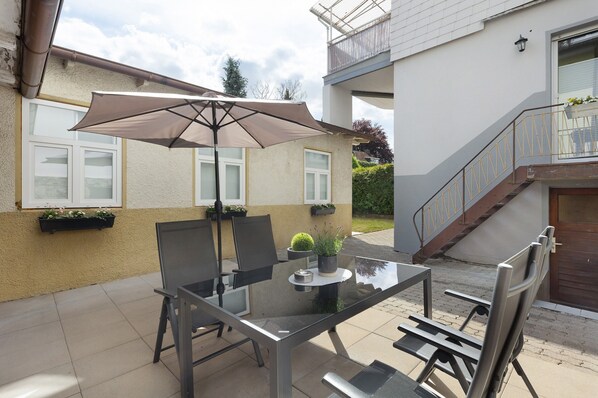Een terras buiten met een glazen tafel, stoelen en een grote parasol.
