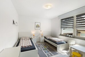 Een kamer met twee bedden, een bureau, een stoel, een raam met zonwering, een plafondlamp en een kinderspelmat met hopscotch.