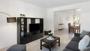 Een moderne woonkamer met een flatscreen televisie, donkere meubels, een kleine tafel met een vaas en een eethoek op de achtergrond.