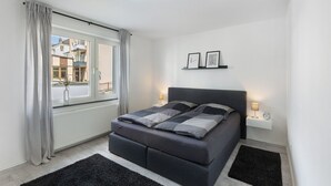 Een moderne slaapkamer met een bed, een raam met gordijnen, een nachtkastje met een lamp, en een wandplank met foto's.