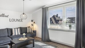 Ein modernes Wohnzimmer mit einer Couch, einem Couchtisch, einer Pendelleuchte und Blick auf Gebäude durch ein Fenster.