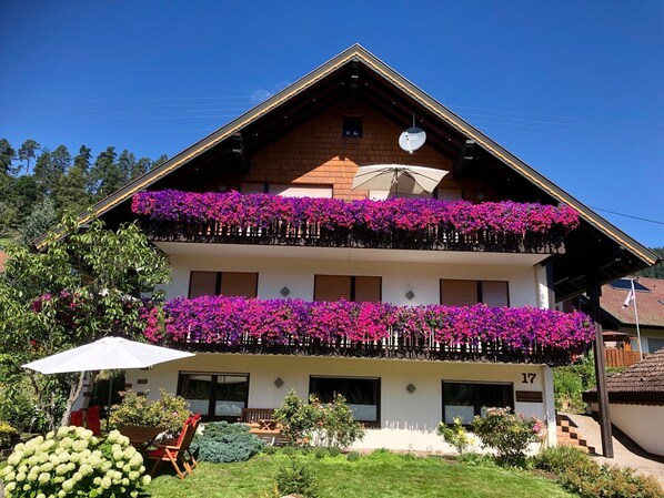 A two-story building with balconies adorned with flowers, a visible number '17', and a garden area with a table and chairs.